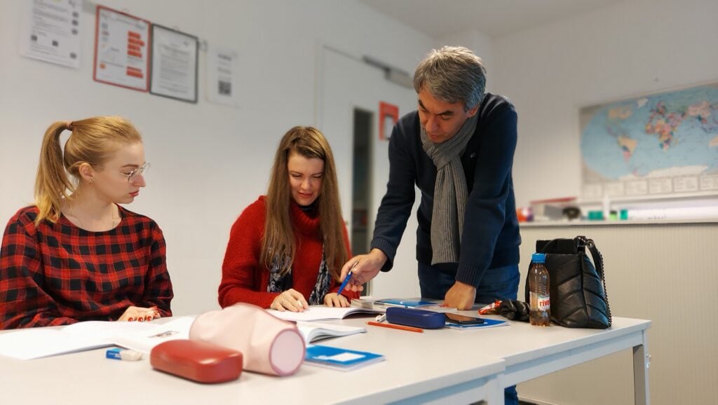 Lehrer erklärt zwei Teilnehmerinnen etwas und zeigt mit einem Stift auf ein offenes Buch auf dem Tisch; beide Frauen folgen aufmerksam seinem Hinweis.