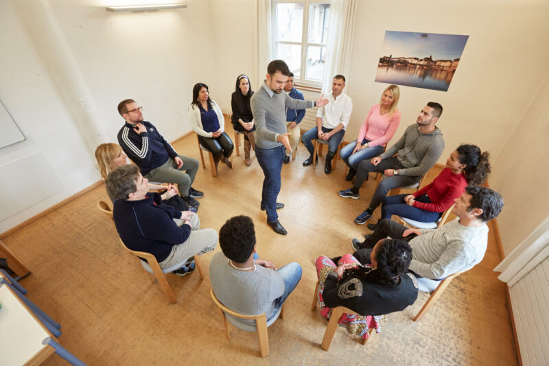 The instructor speaks to a group of students sitting in a circle of chairs